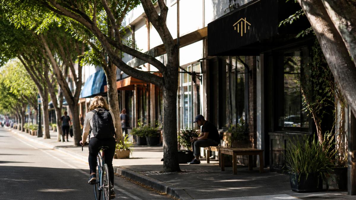 A cyclist riding along a tree-lined downtown Ventura street with local shops and cafes visible, representing walkable commute options