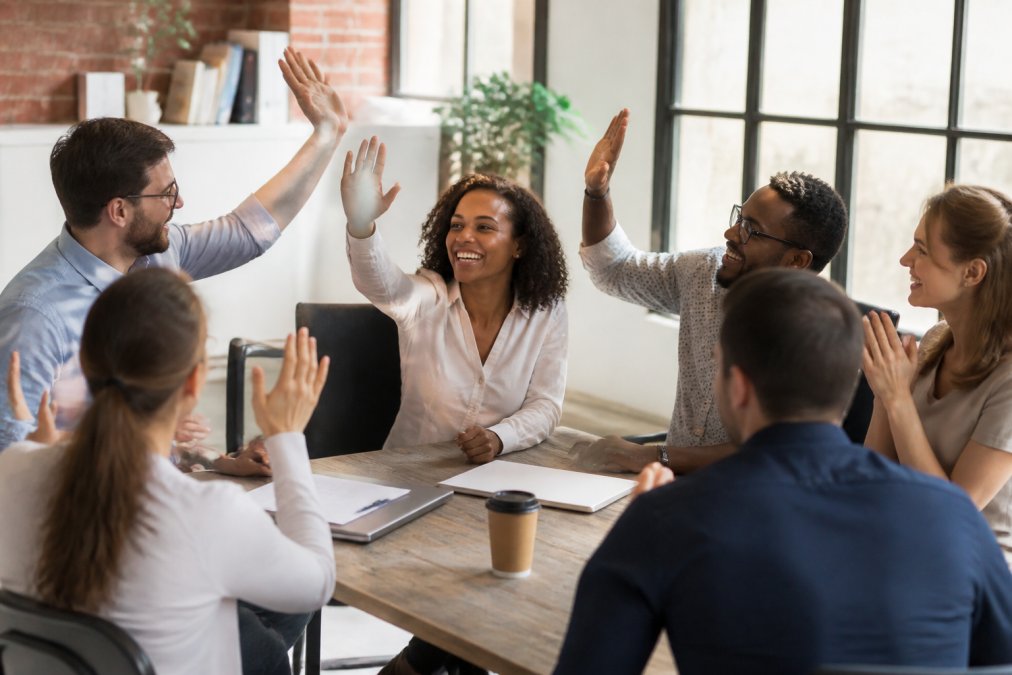 Small team of diverse employees gathered around a conference table, smiling and giving high-fives to celebrate a colleague's achievement, natural office lighting streaming through windows