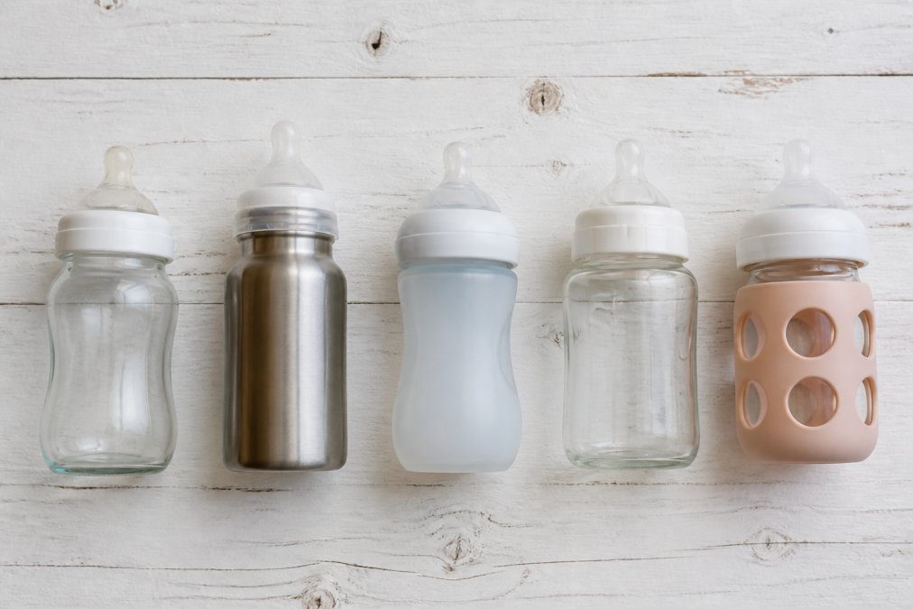Flat-lay of five baby bottles in different materials, clear borosilicate glass, brushed stainless steel, soft silicone, wide-neck glass, and a hybrid bottle with silicone sleeve, arranged neatly on a clean white wooden surface with soft natural light