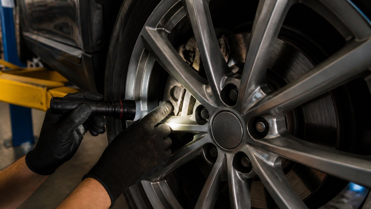 Close-up of a mechanic's gloved hands using a flashlight to inspect a brake pad through the wheel spokes of a car on a lift in a clean auto service bay