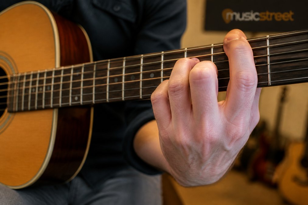 Close-up of a guitarist's fretting hand pressing down hard on acoustic guitar strings, knuckles whitening, fretboard and worn frets clearly visible under warm indoor lighting