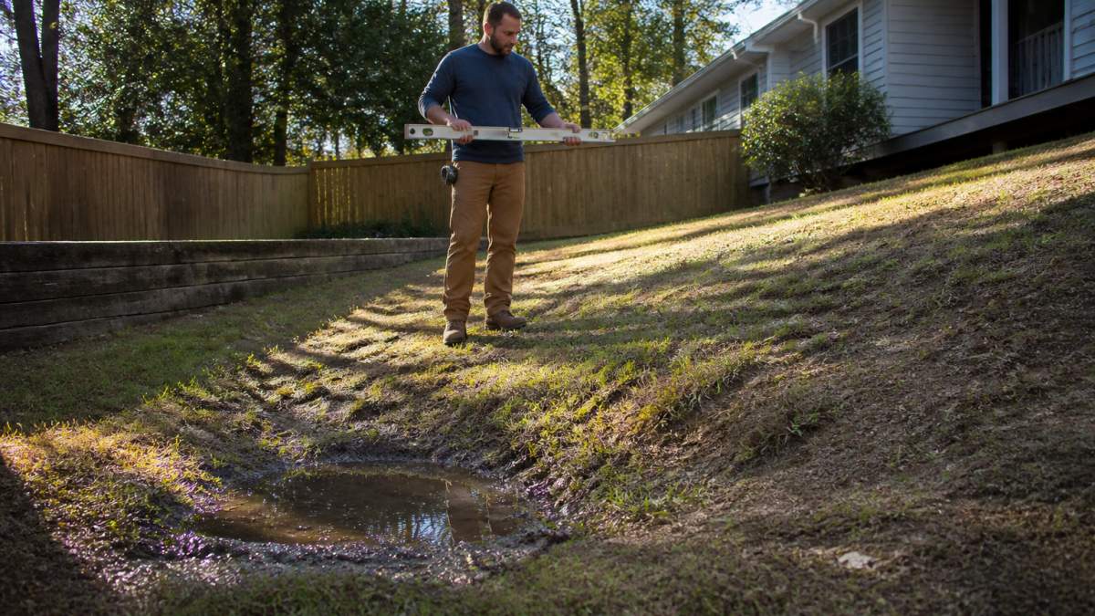 Homeowner standing in a sloped backyard holding a long level tool, examining visible low spots and uneven terrain with water pooling in a depression, morning sunlight casting shadows across the uneven ground