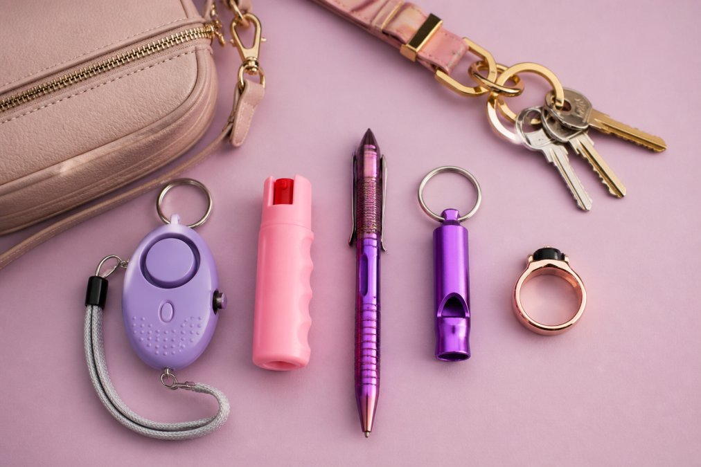 Flat lay of colorful self-defense accessories, a personal alarm, a small pepper spray canister, a tactical pen, a safety whistle, and a wearable ring, arranged neatly on a pastel background alongside a set of keys and a small crossbody bag