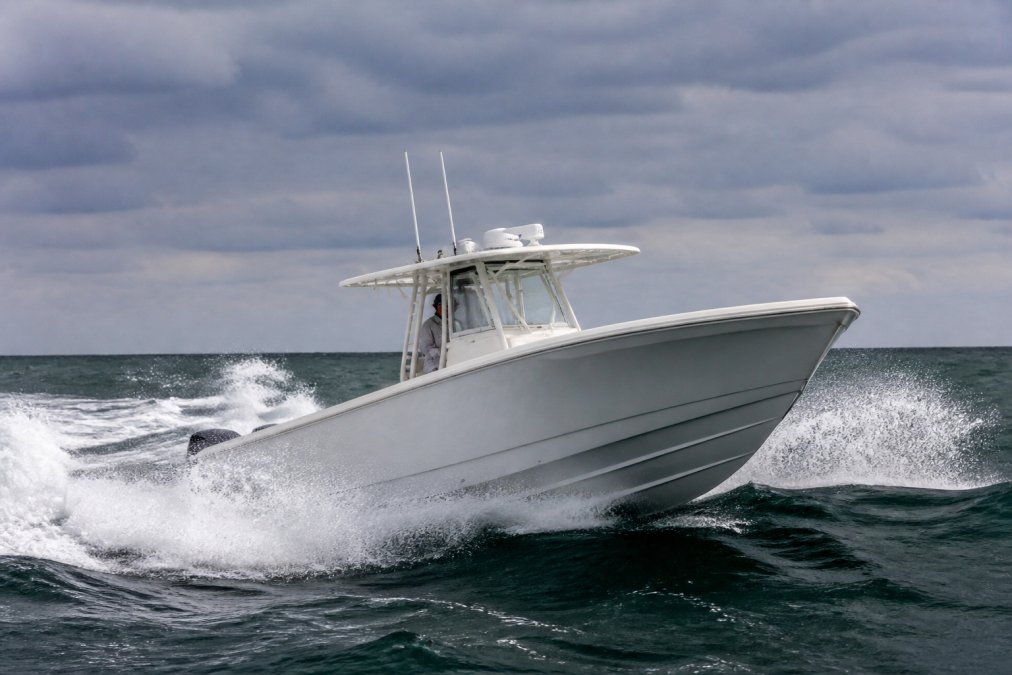 A center console boat cutting through choppy offshore waters, deep-V hull entry and bow flare visible as spray deflects away from the cockpit, overcast sky and dark green ocean