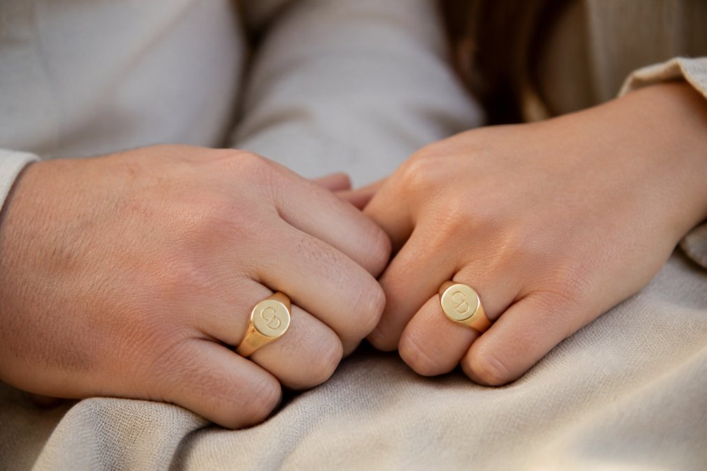 Close-up of two hands wearing matching gold signet rings with engraved initials, resting on a soft cloth in warm natural light