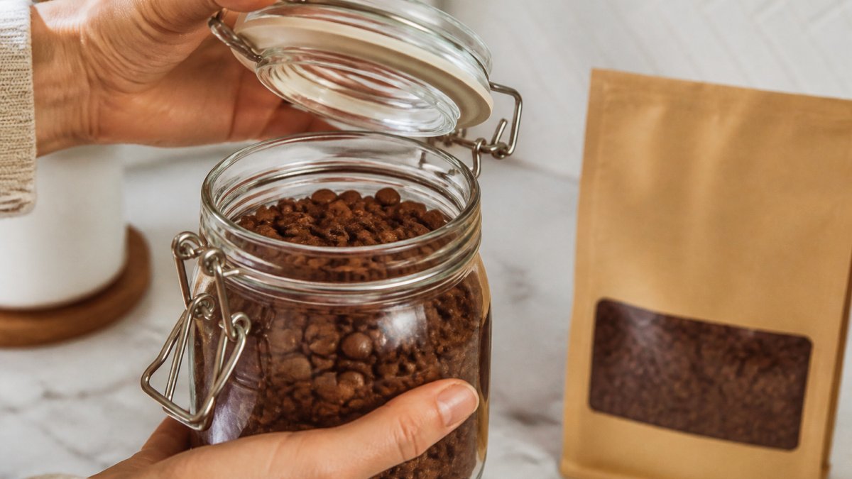 Close-up of hands opening an airtight glass coffee canister with whole beans visible inside, showing the rubber seal mechanism and a Tinto coffee bag in the background on a kitchen counter