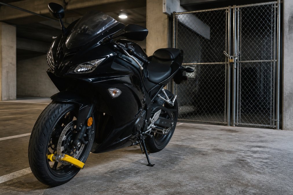 Motorcycle secured with heavy-duty alarm disc lock attached to front wheel, parked in covered garage with metal storage cage visible in background, secured with chain