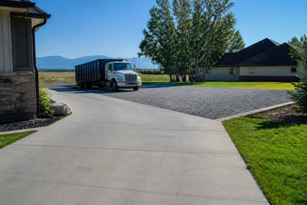 A residential driveway with a clear path leading to a level gravel pad, with a roll-off dumpster truck visible in the background approaching the drop-off zone under bright morning light