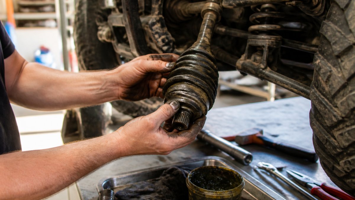 Mechanic's hands inspecting a CV axle boot for tears and checking grease condition on an off-road vehicle in bright shop lighting