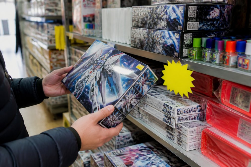 Close-up of hands holding a model kit box while browsing a retail clearance shelf stacked with hobby supplies and discounted items, fluorescent store lighting overhead