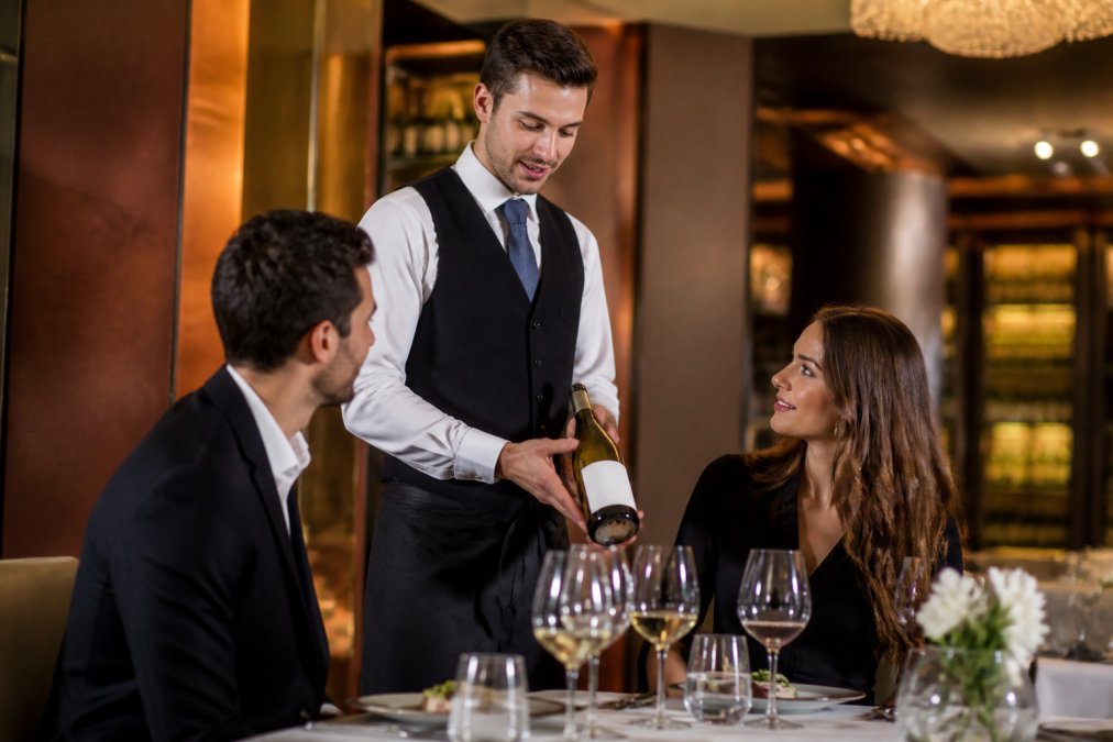 A professional hotel server in a smart uniform presenting a bottle of white wine to guests at a table in an upscale restaurant setting with warm ambient lighting