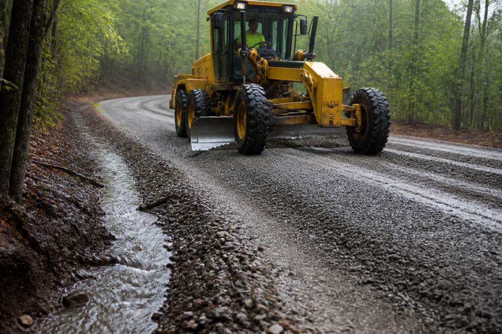 A professional operator using a motor grader to shape a crowned gravel driveway, with water visibly running off the side into a shallow ditch during a light rain