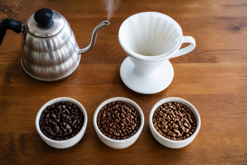 A clean overhead shot of a white pour over dripper on a wooden counter, a gooseneck kettle steaming beside it, and three small bowls of whole roasted coffee beans in different shades of brown