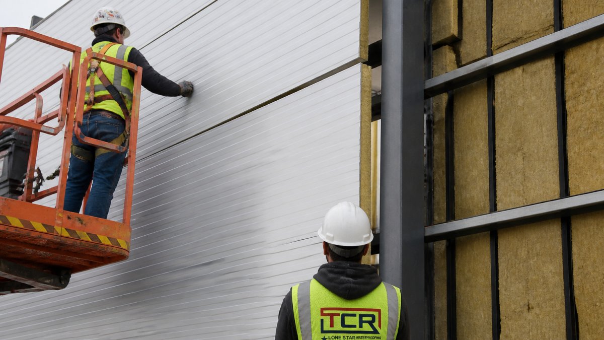 Close-up of a crew installing large insulated metal wall panels with a lift on an industrial warehouse, showing the panel tongue-and-groove joint and concealed fasteners