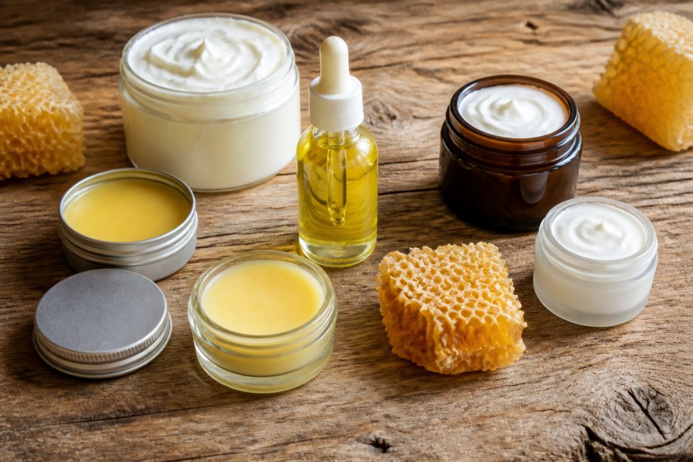 A flat lay of six distinct natural moisturizer products including a jar of whipped body butter, a glass dropper bottle of facial oil, and a tin of balm, arranged on a rustic wooden table with soft natural light and a few raw honeycomb pieces nearby