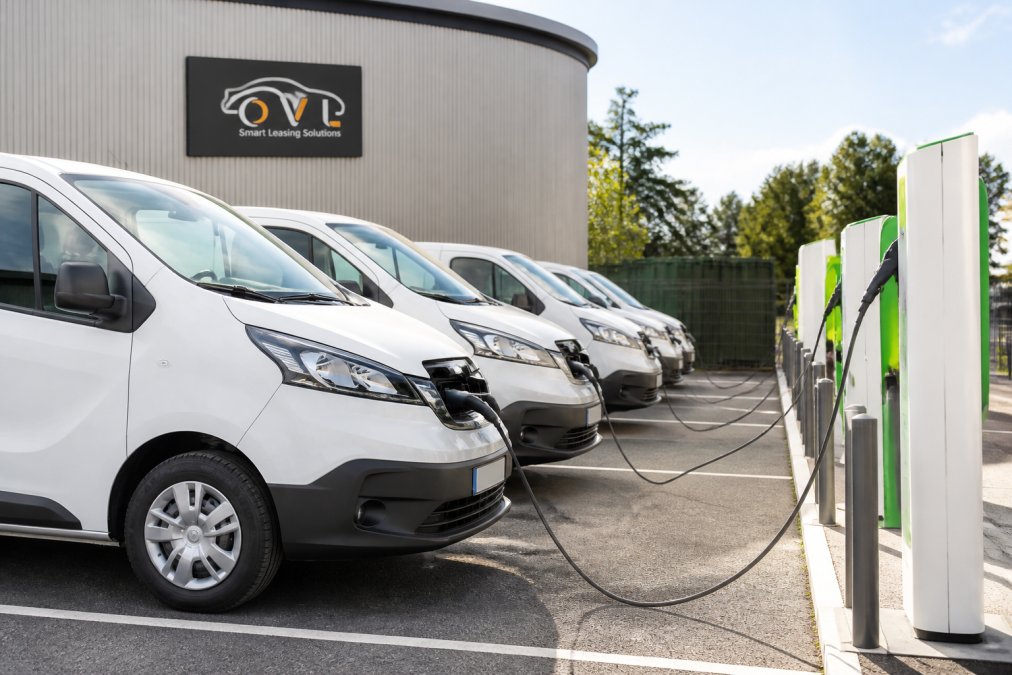 Fleet of modern electric vans parked in a row at a charging station with visible charging cables and green energy infrastructure