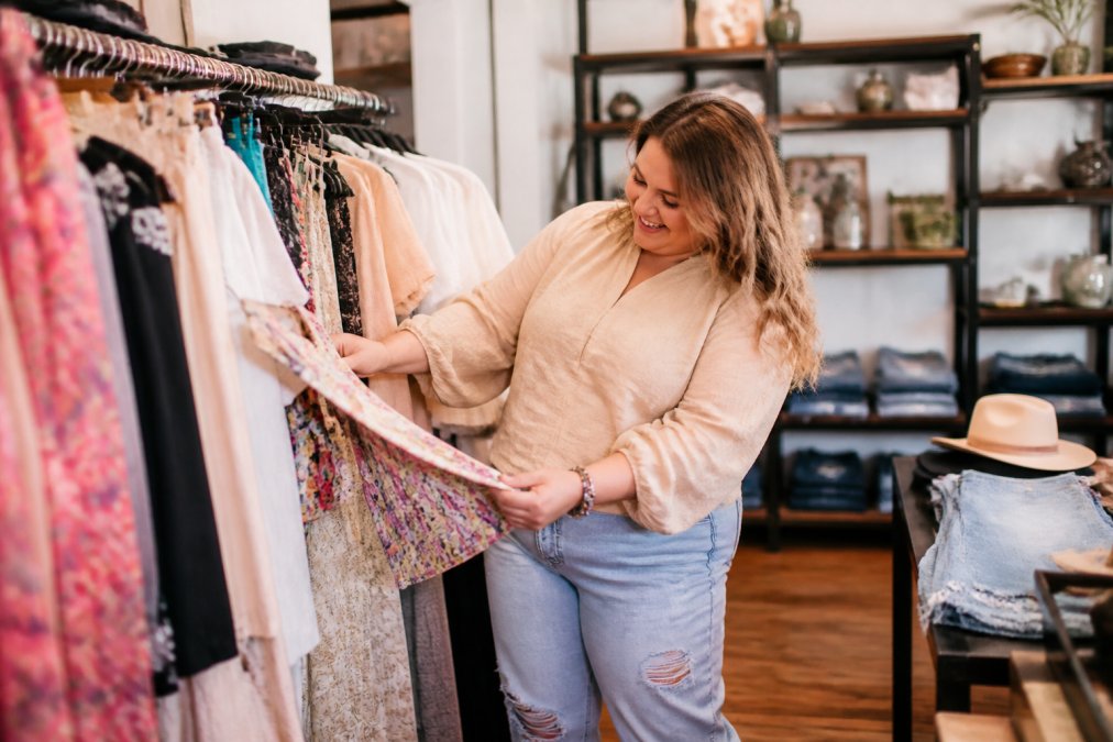 Woman with a curvy silhouette smiling while browsing colorful clothing on display racks in a bright, welcoming boutique setting with warm lighting and carefully curated pieces