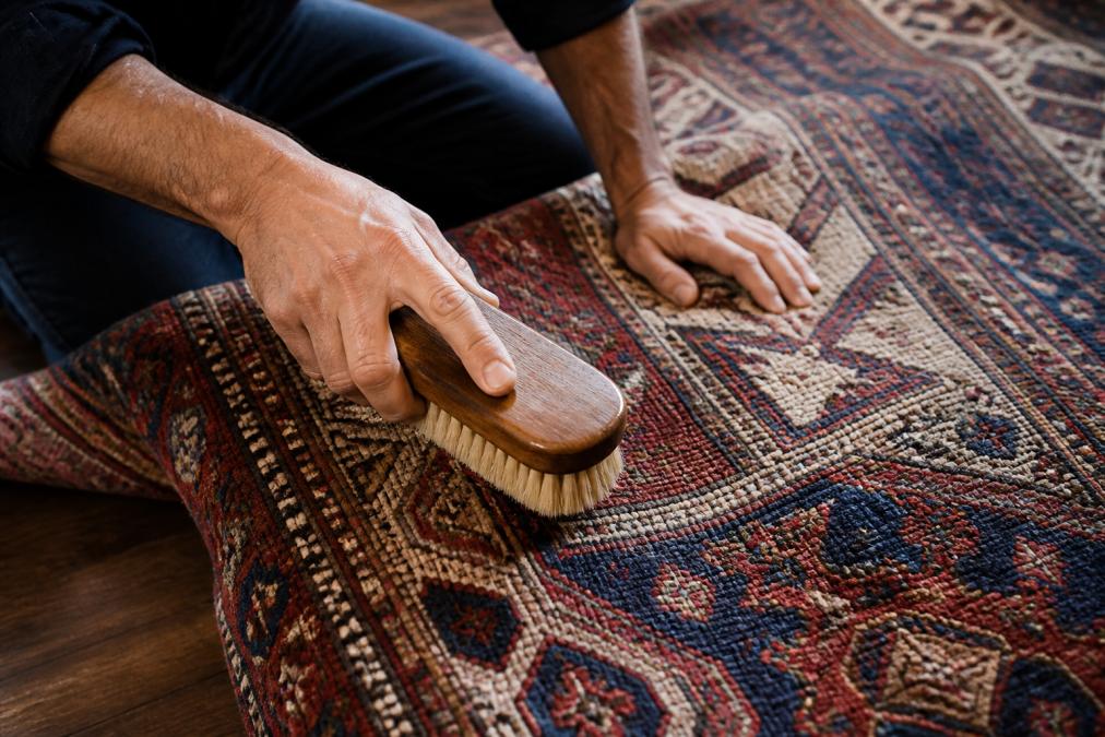Close-up of a person's hands gently brushing a hand-knotted wool rug with a soft-bristle brush, revealing the intricate weave and natural fiber texture in warm natural light