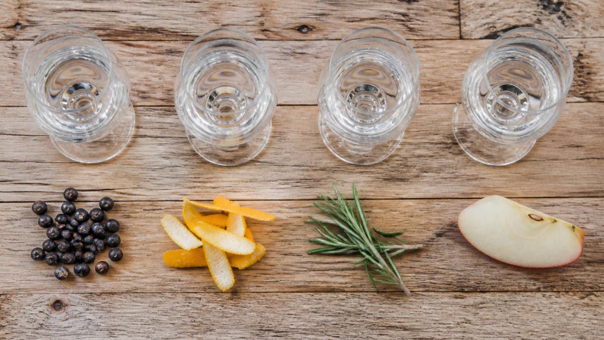 Overhead shot of four small gin tasting glasses on a rustic wooden table, each with a different garnish beside it: juniper berries, citrus peel, a sprig of rosemary and a slice of apple