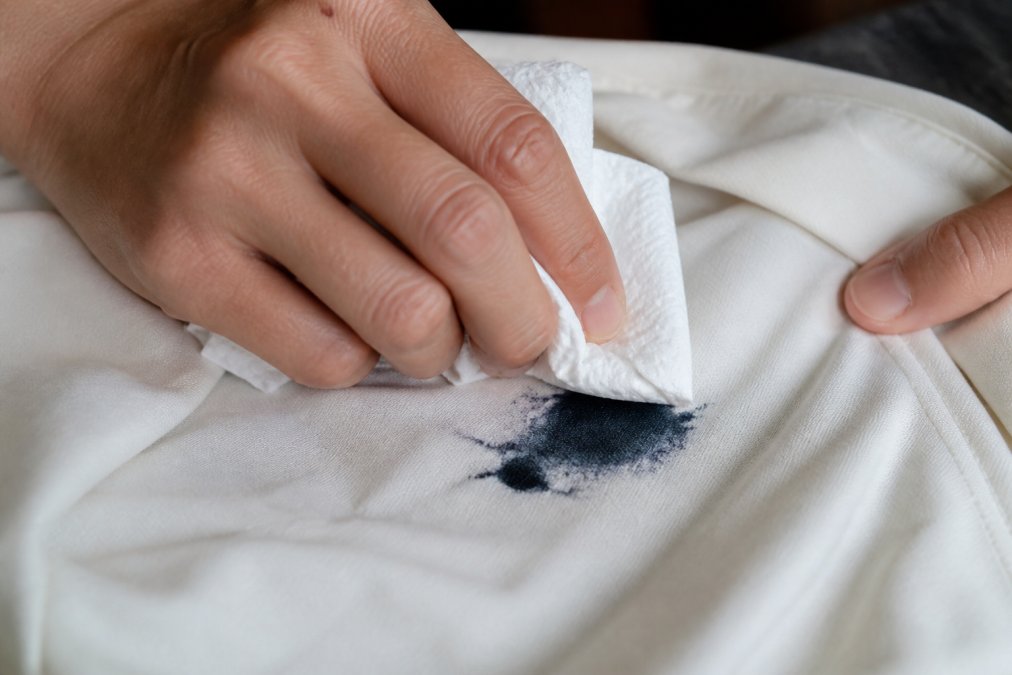 Close-up of hands using white paper towels to blot a fresh ink stain on polyester fabric, showing the dabbing motion without rubbing, with natural light highlighting the stain