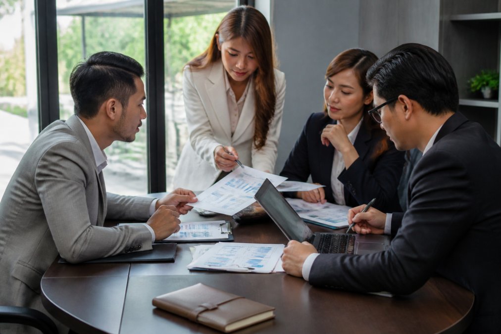 Team of business professionals analyzing financial reports and profit margins together at conference table with laptops and spreadsheets, morning natural light