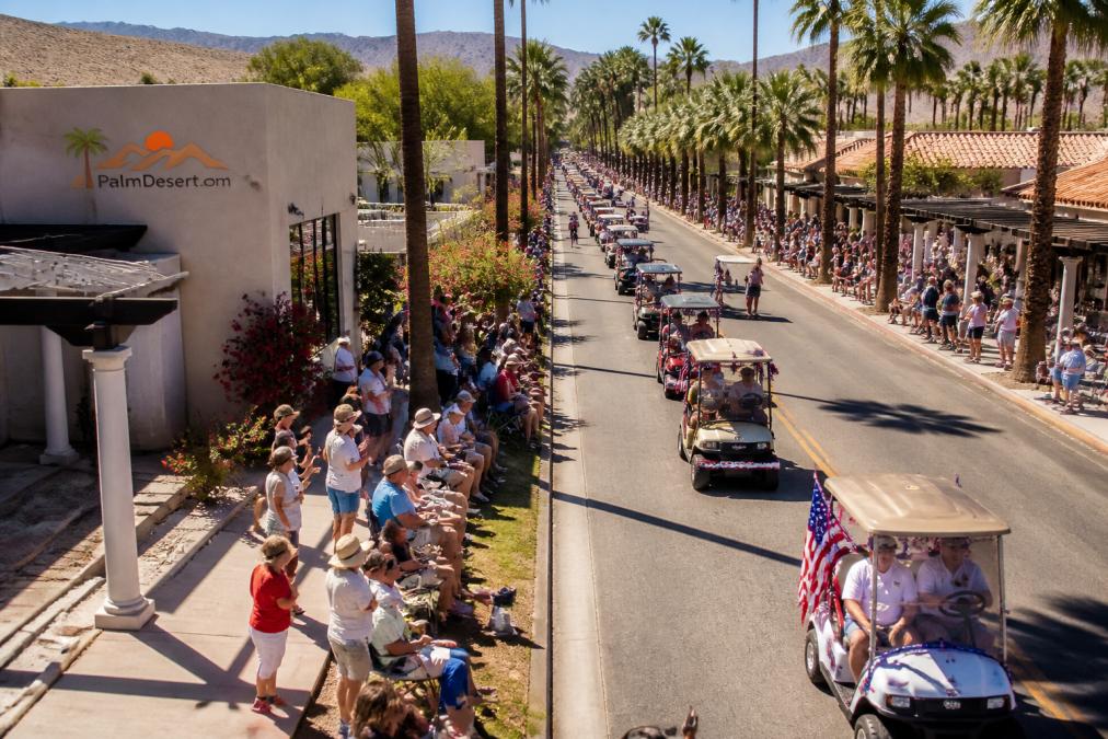 Crowds watch as decorated golf carts move down the sunny, palm-lined street during a festive golf cart parade.