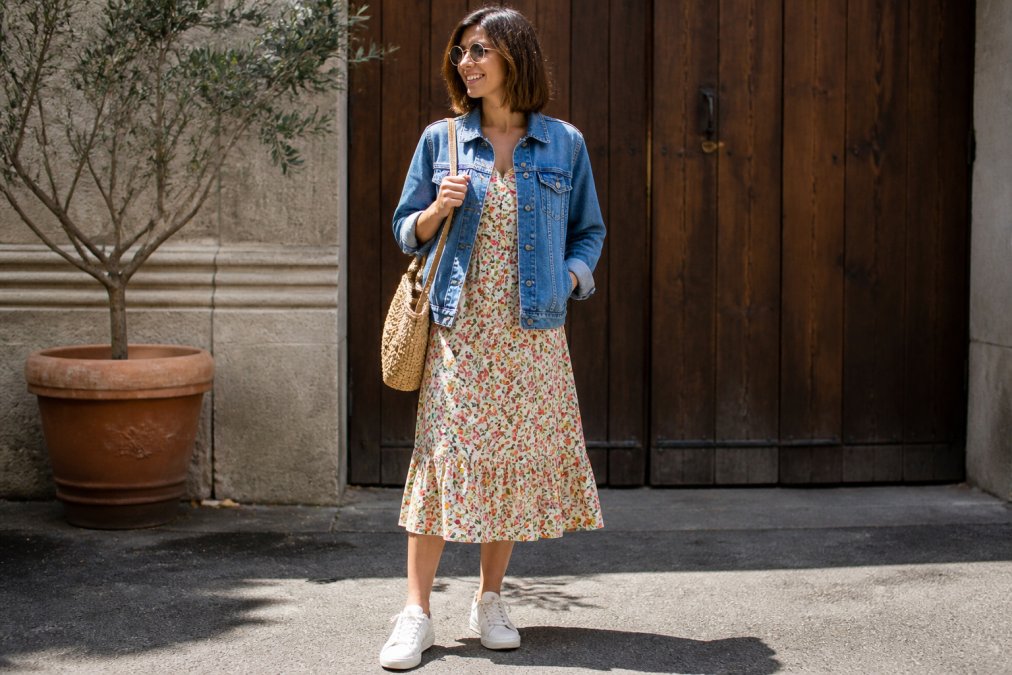 Woman wearing a vintage-inspired floral dress styled with modern white sneakers and a contemporary denim jacket, standing in natural daylight