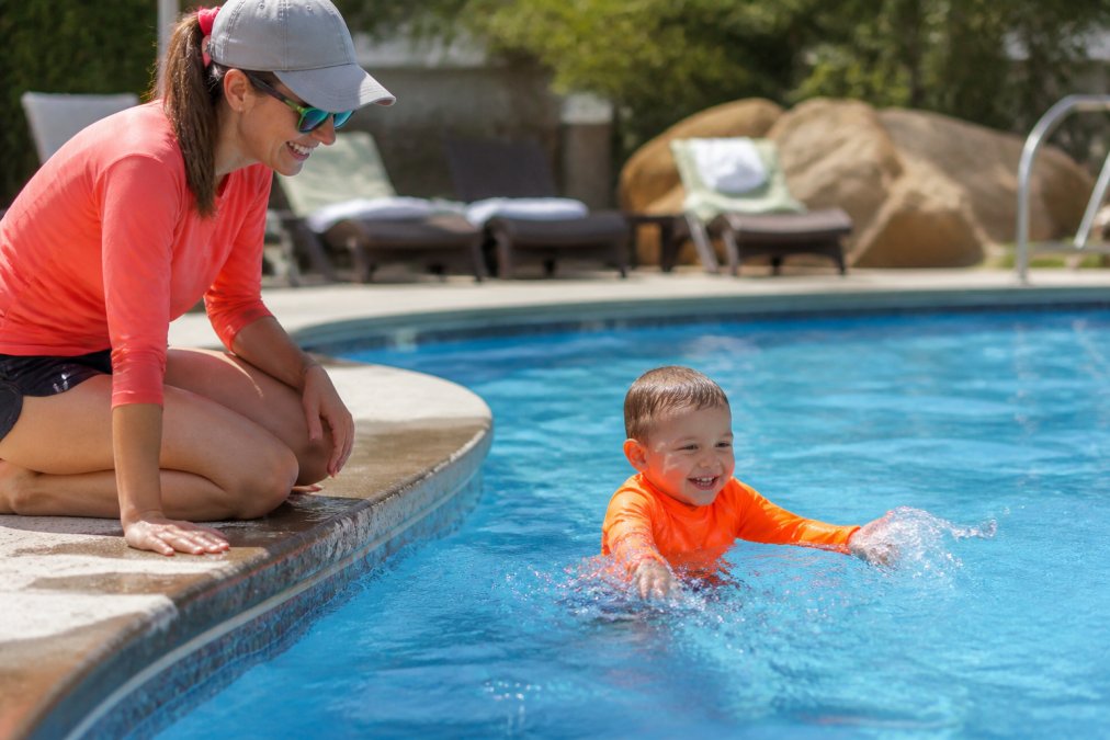 A parent kneeling at the edge of a sunlit pool watching a toddler in a bright neon orange rash guard splashing in clear blue water, the child's vivid color contrasting sharply with the water