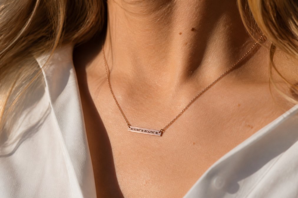 Woman wearing a delicate personalized necklace with custom coordinates pendant in rose gold, natural sunlight illuminating the intricate metalwork against her collarbone