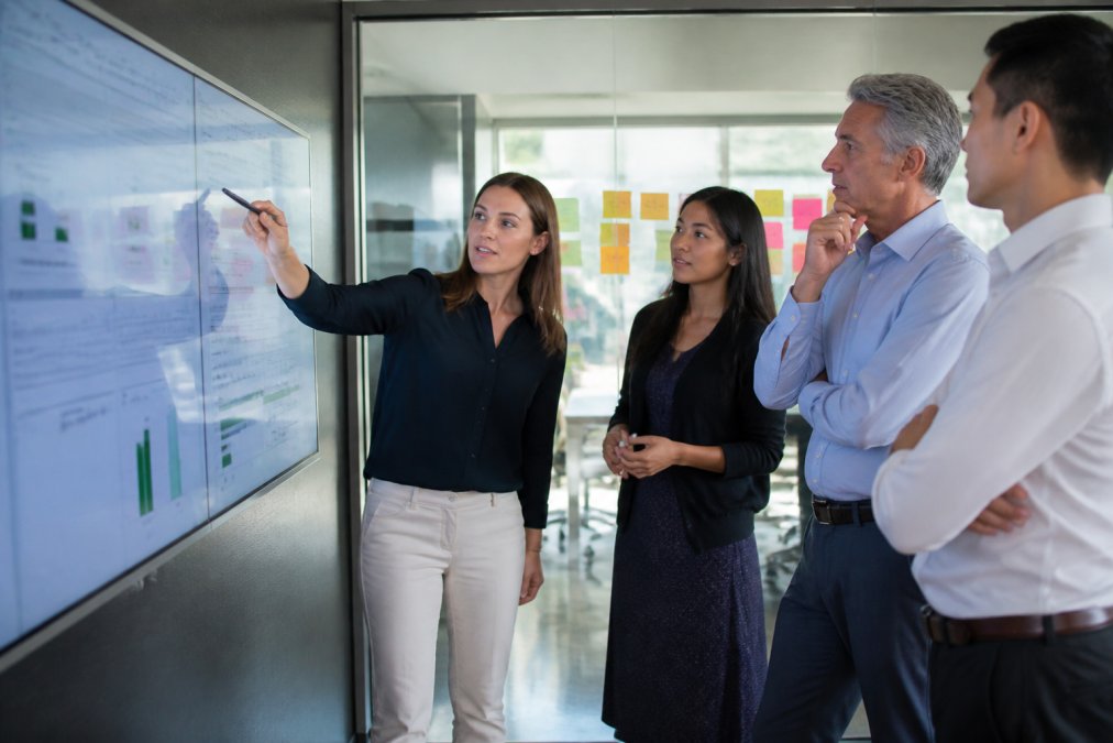 Business team gathered around a wall-mounted screen reviewing quarterly initiative progress with sticky notes on a glass board