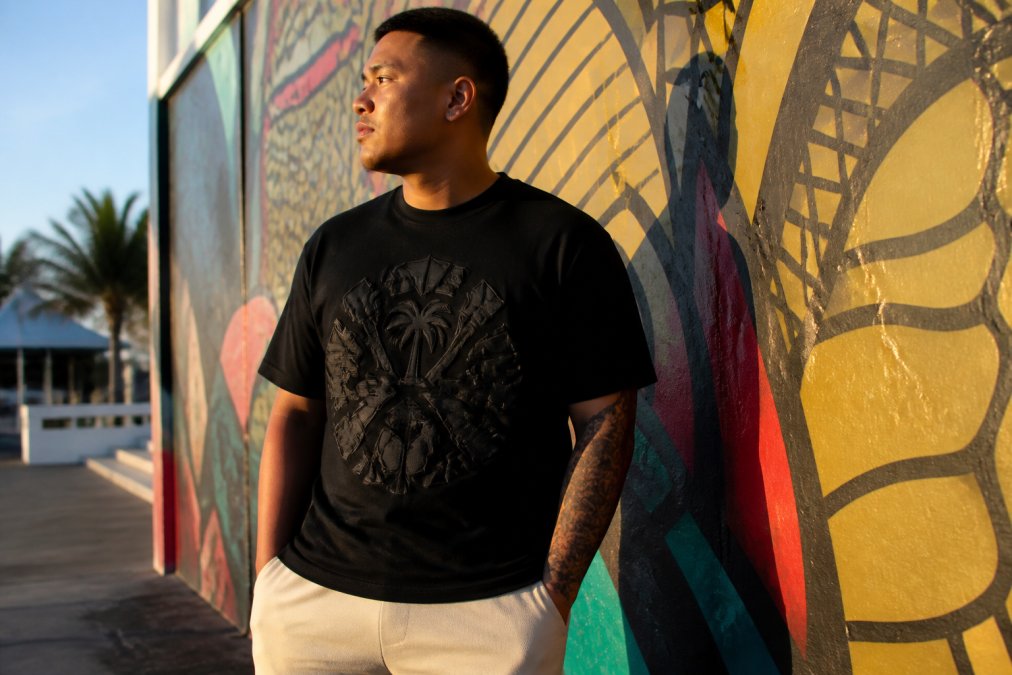 A young Chamorro man in a black puff-print graphic tee standing confidently against a colorful urban mural, golden hour lighting casting long shadows