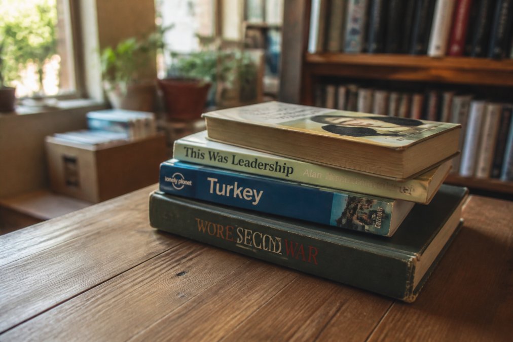 A stack of well-loved secondhand books, including a Lonely Planet travel guide and a military history title, resting on a wooden table in a cozy bookshop with warm afternoon light streaming through a window