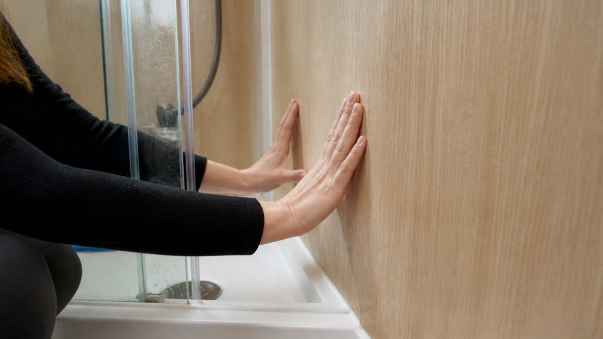Close-up of hands running along a wood-effect wall panel beside a shower enclosure in a small modern bathroom, water droplets on the glass, sealant line visible at the base