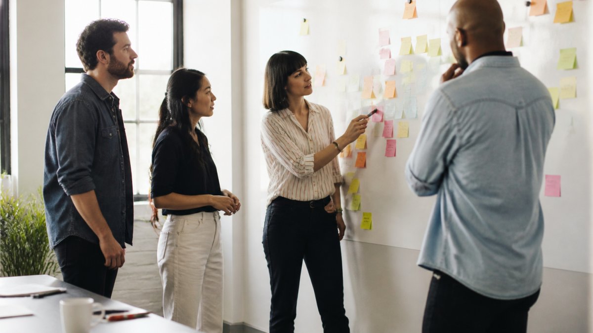 A digital agency team gathered around a whiteboard covered in sticky notes, discussing sprint priorities with a client in a bright modern office