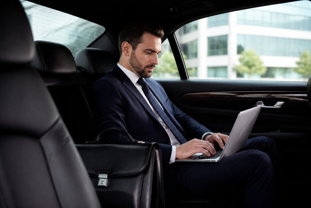 A business executive in a tailored suit working on a laptop in the quiet backseat of a luxury sedan, leather briefcase beside them, soft daylight through tinted windows