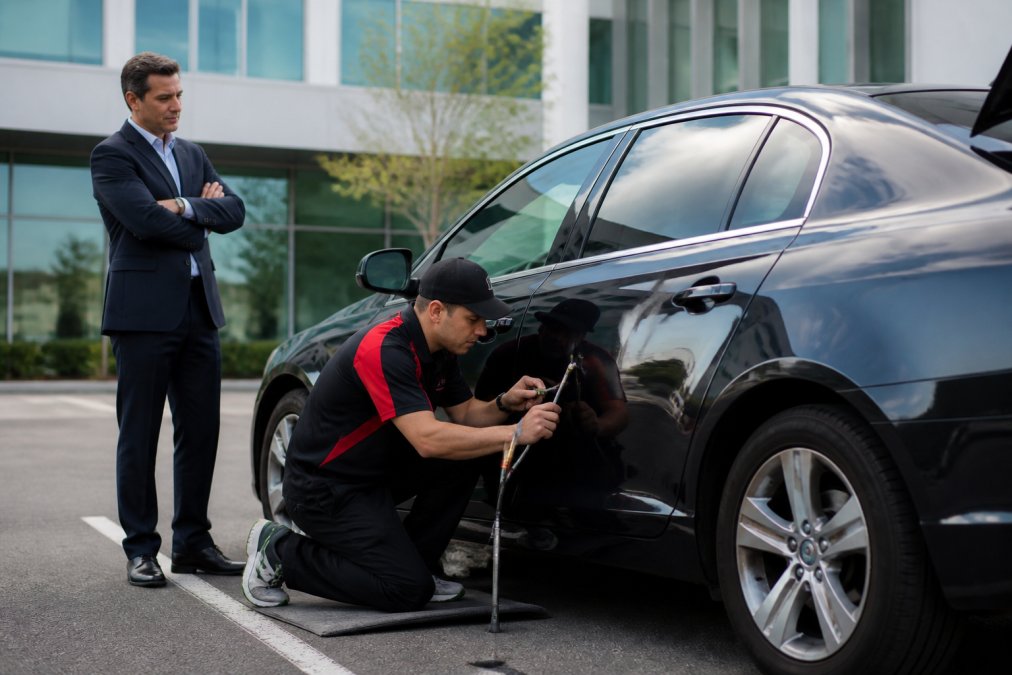 Professional in business attire standing beside their car in a corporate parking lot while a technician uses specialized PDR tools to repair a door ding on the vehicle
