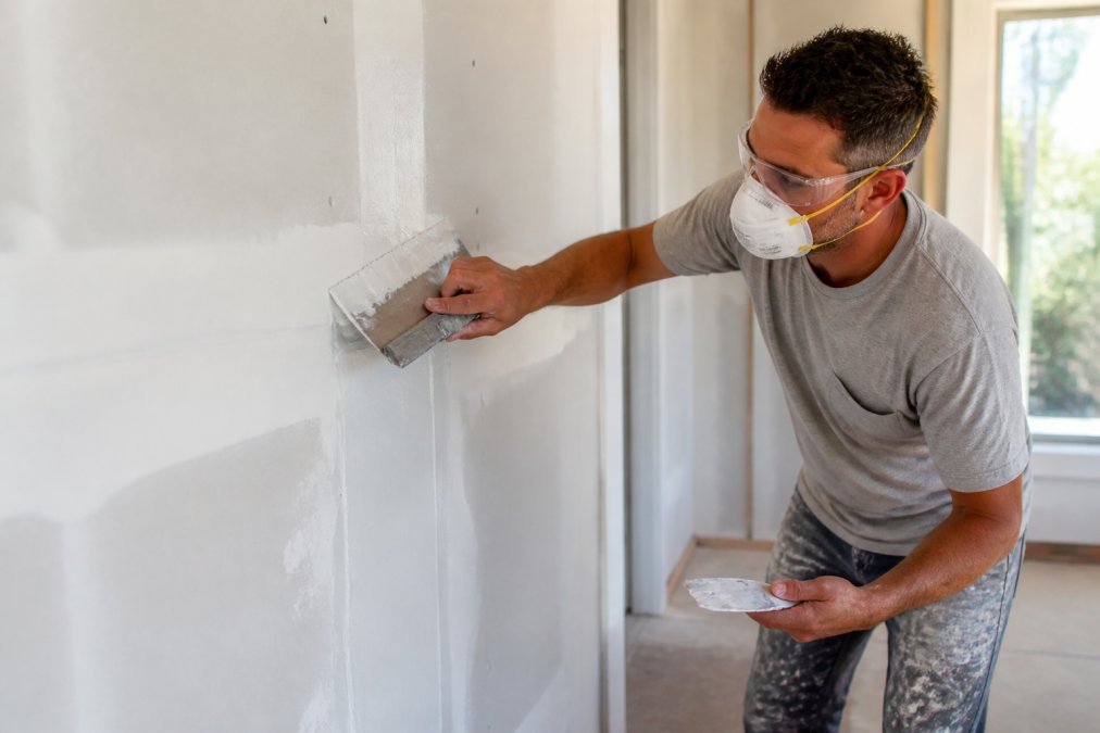 A professional drywall finisher using a wide taping knife to apply joint compound over a seam, with a dust mask and safety glasses nearby, natural window light across the wall
