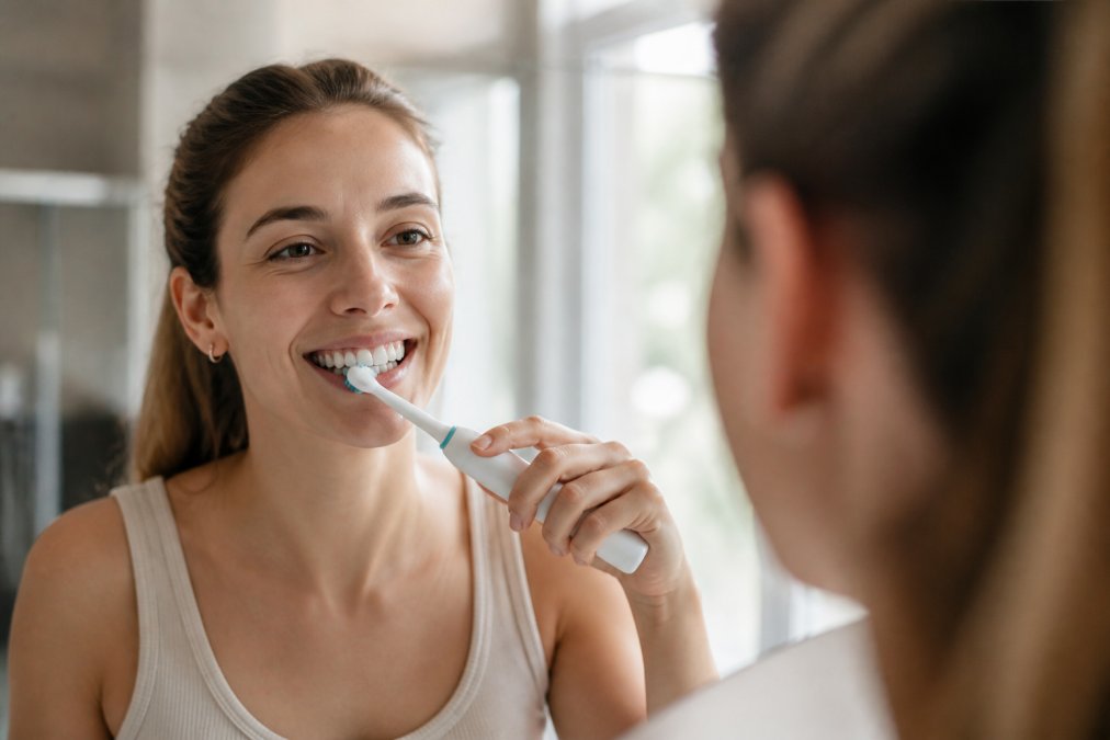 Close-up of a person using an electric toothbrush on straight teeth, bright bathroom mirror and natural window light behind them