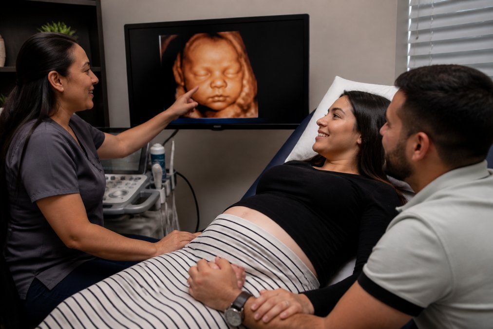 Expectant parents smiling as they view their baby's 3D ultrasound image on a large screen, technician pointing to facial features while the mother holds her partner's hand in a comfortable clinical setting with soft lighting
