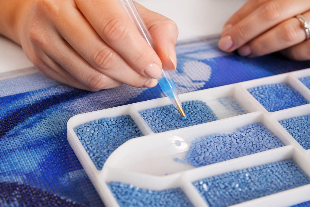 Close-up of a crafter's hands using a diamond painting pen loaded with blue putty, picking up a resin drill from a divided plastic tray under bright desk lighting
