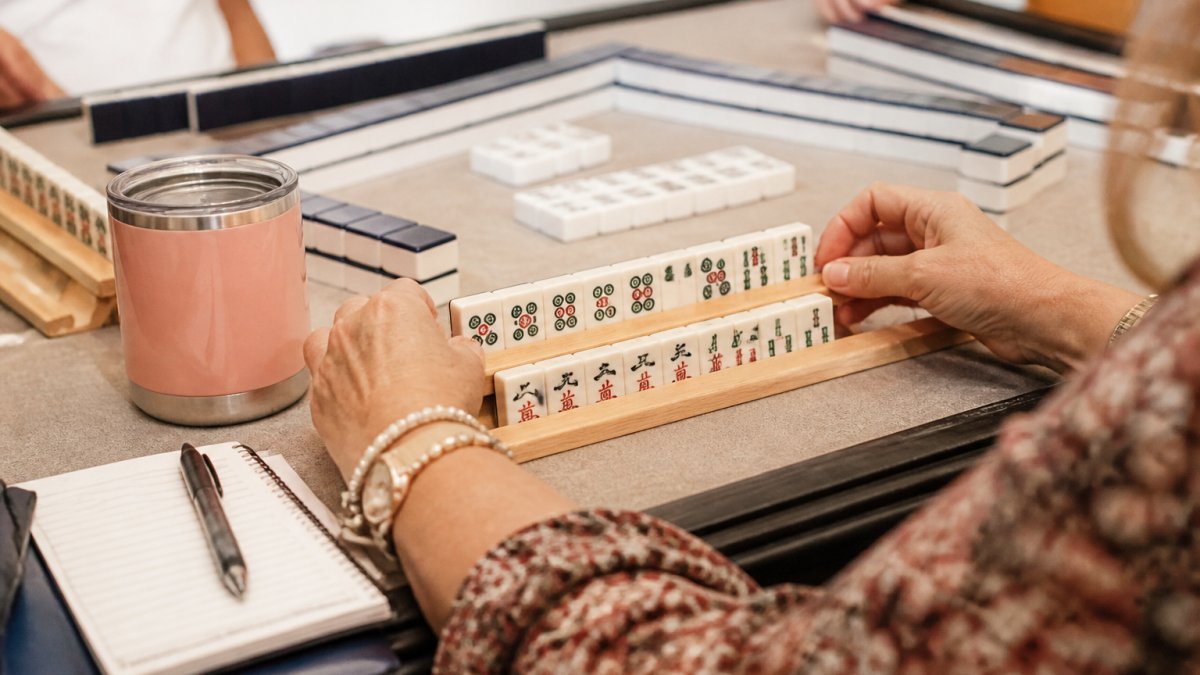 Close-up of a woman's hands arranging Mahjong tiles on a two-row wooden rack at a table set for game night, with a game mat underneath and other players' racks visible in soft focus