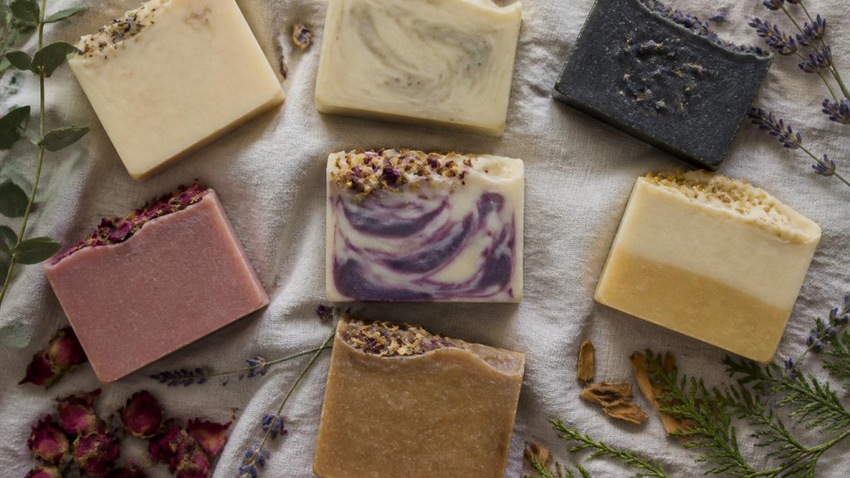 Overhead flat lay of seven colorful handcrafted soap bars arranged artfully on a natural linen cloth with fresh botanicals including lavender sprigs, rose petals, and cedarwood pieces scattered around them