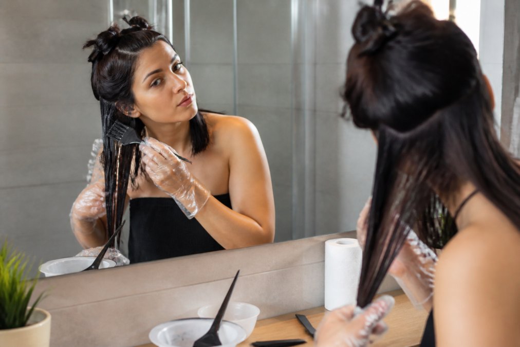 Woman wearing gloves applying at-home keratin treatment to sectioned hair with a tinting brush in a bright bathroom