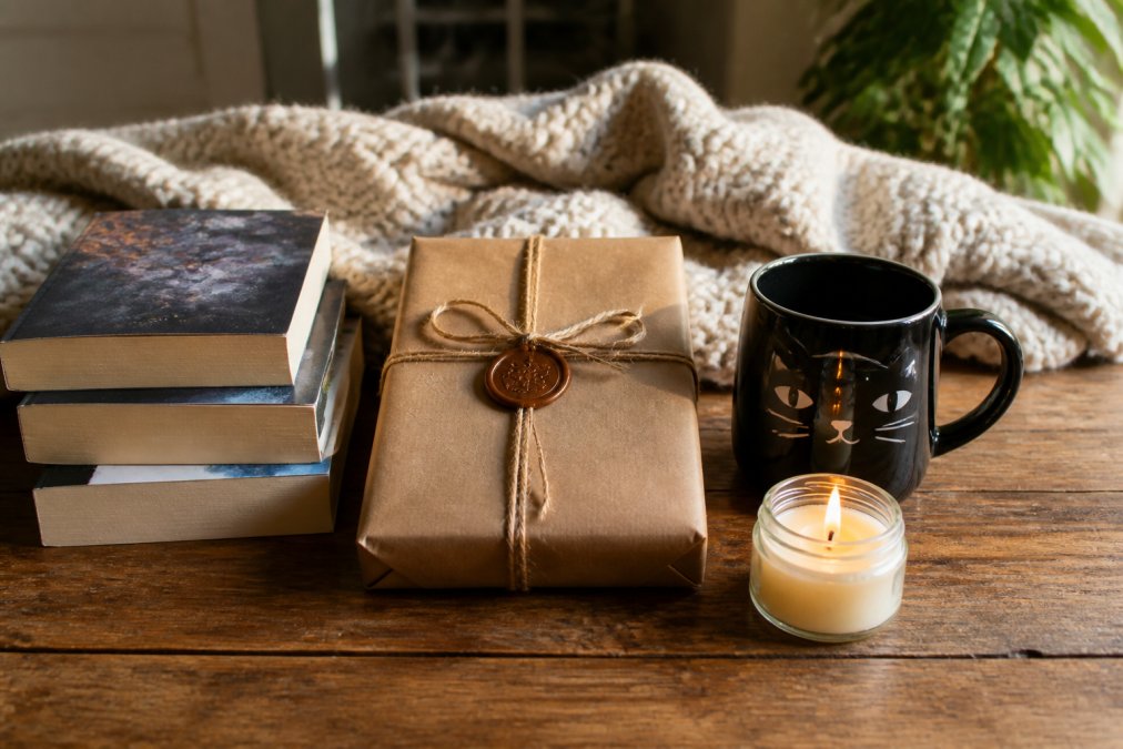 A flat lay of cozy mystery book gift sets on a warm wooden table, featuring a wrapped mystery book, a glossy black cat mug, a small lit soy candle, and a stack of well-loved paperbacks, styled with soft afternoon lighting and a knitted blanket in the background