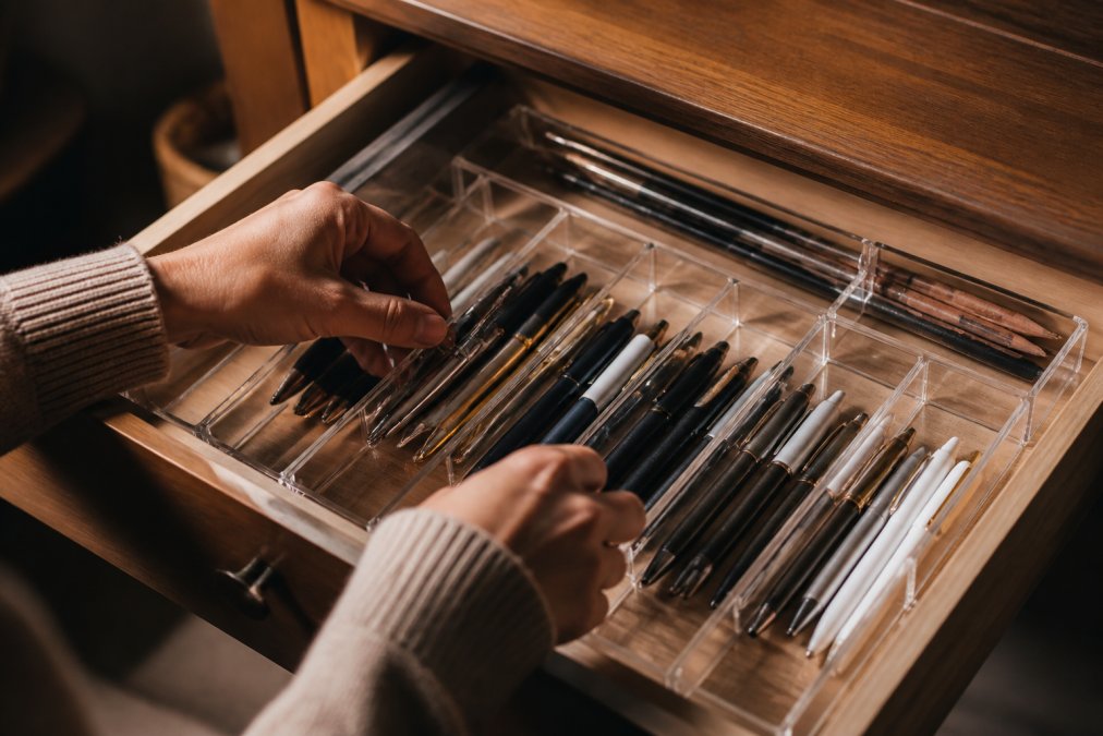 Close-up of hands arranging multiple writing instruments and pens neatly into a clear acrylic organizer tray inside an open desk drawer, showing compartments and dividers in use