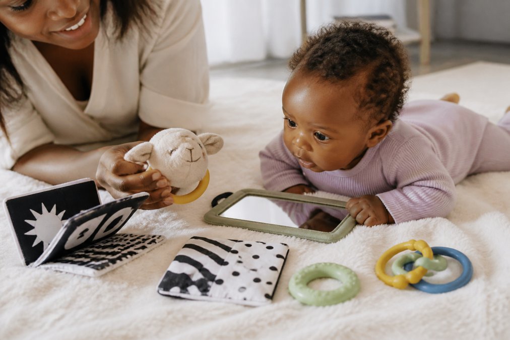 A flat lay photograph of seven newborn developmental toys, including a black-and-white contrast book, a soft plush rattle, a small tummy time mirror, a crinkle fabric square, a silicone teether, and a set of grasping rings, arranged neatly on a soft white blanket in bright, natural daylight