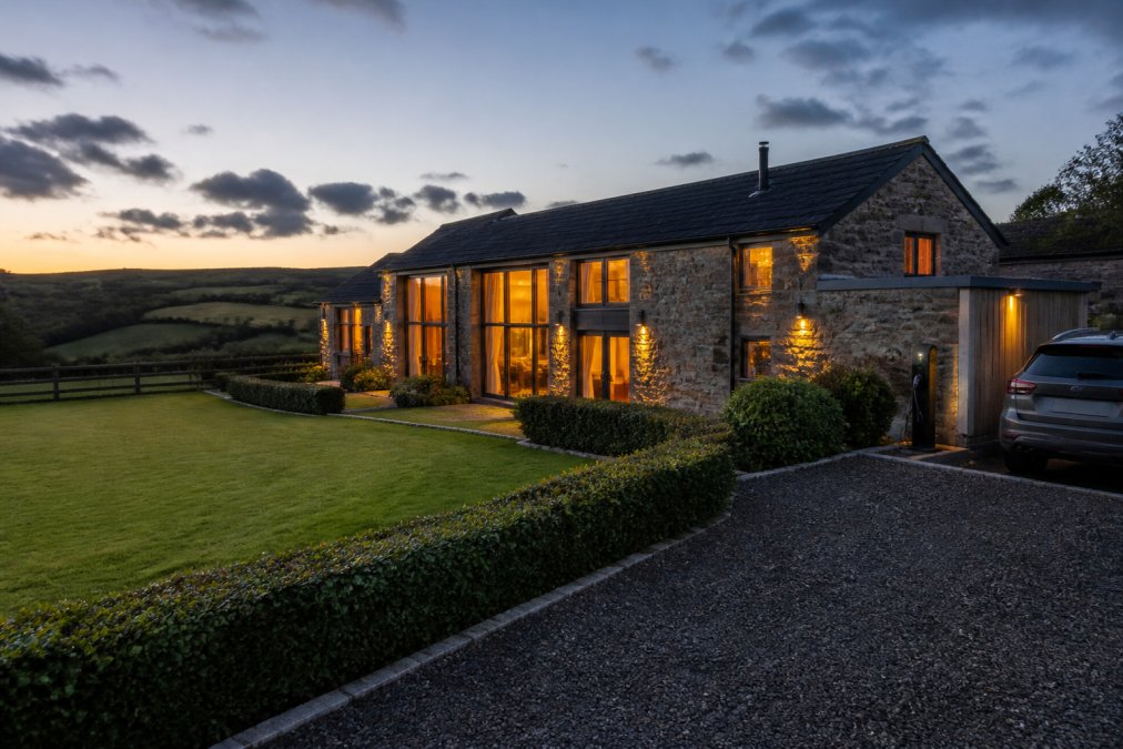Modern barn conversion with warm evening lighting glowing from large windows, stone exterior walls, manicured garden with green hedging, and countryside rolling hills visible in background at dusk