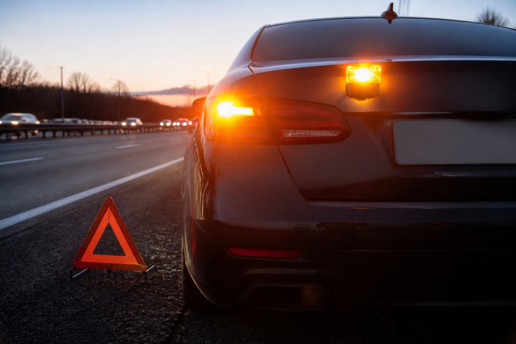 7 Best Emergency Beacons for Car Breakdowns 2026 A close-up, eye-level photograph of a car's rear bumper at dusk on the hard shoulder of a motorway. A magnetic LED emergency beacon is attached to the boot lid, emitting a bright amber flashing light. The car's hazard lights are also on, and a reflective warning triangle is visible on the ground behind the vehicle.