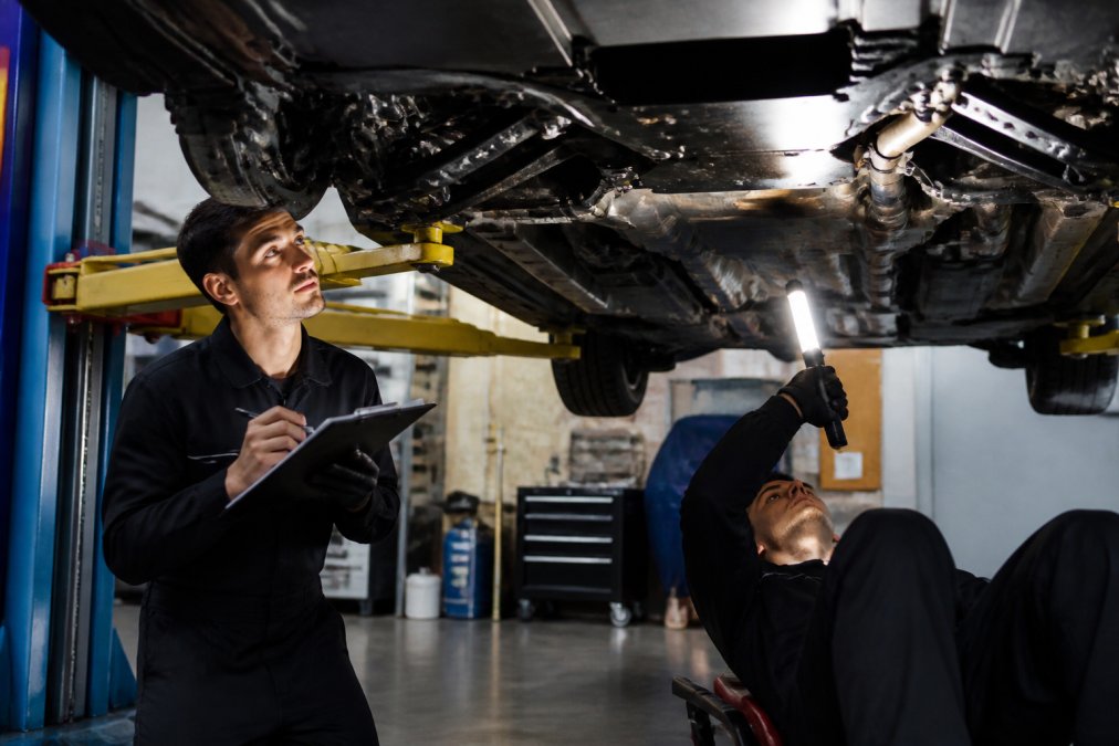 Professional technician performing detailed vehicle inspection on a luxury exotic car, checking undercarriage with bright work lights, documenting findings on clipboard while photographing specific areas