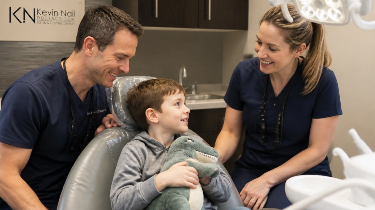 Young child sitting in dental chair with dentist and hygienist nearby, both smiling and making eye contact, with modern dental equipment and bright overhead lighting visible in a welcoming office